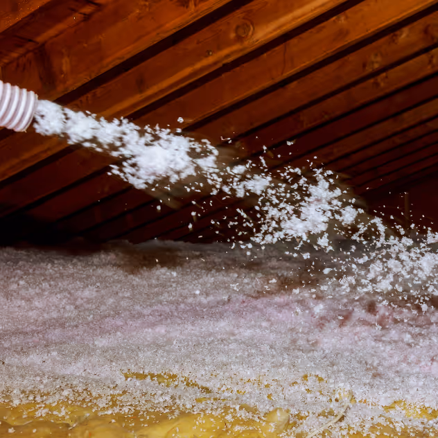 Blown-in insulation being sprayed over existing attic insulation between wooden roof rafters