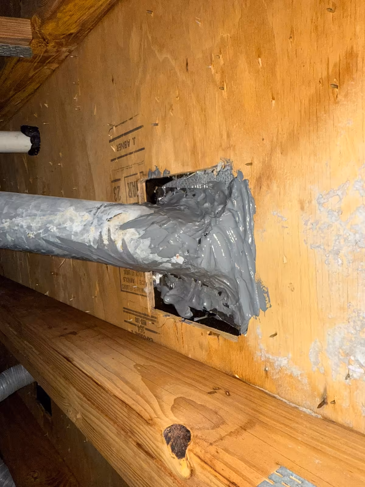 Dryer duct in an attic sealed with grey mastic where it passes through a plywood partition wall