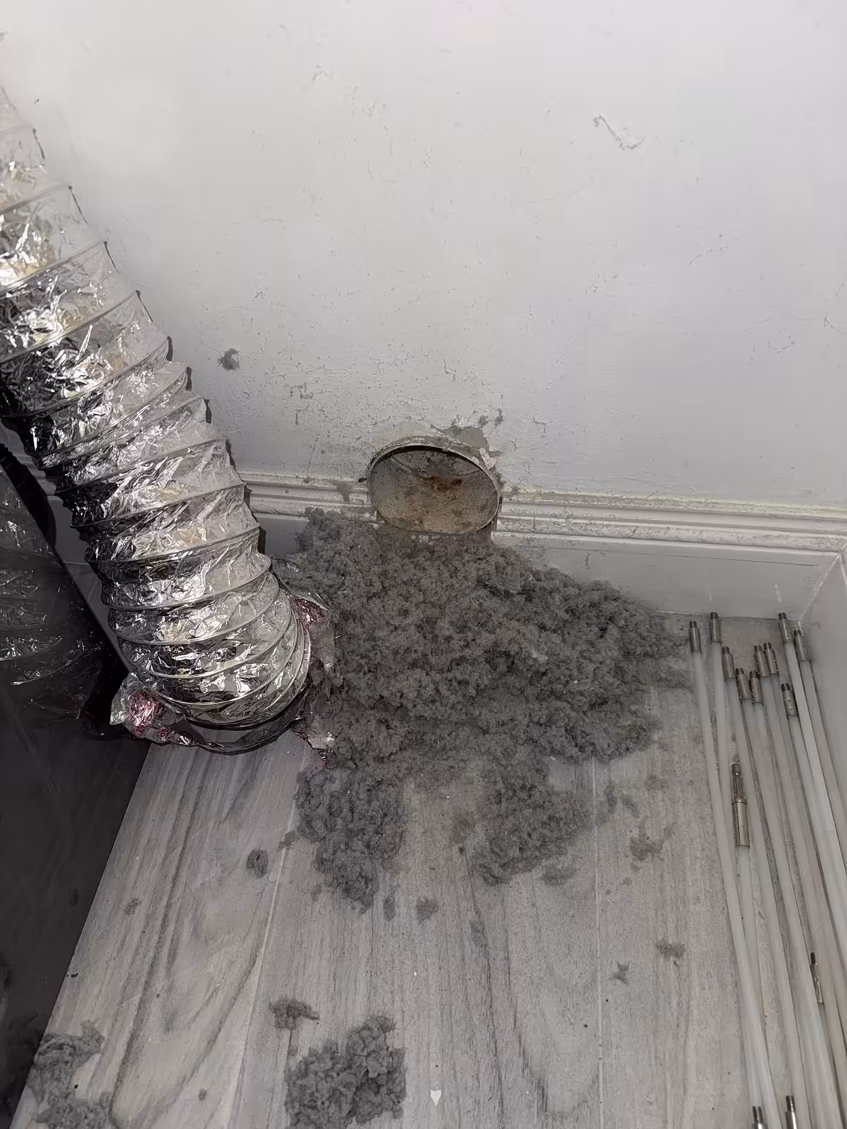 Lint pulled from a round wall dryer vent stub, heaped on the plank floor beside the foil duct and the cleaning rods