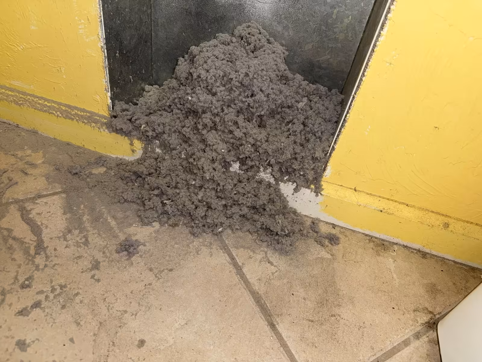 Mound of grey lint spilling out of a recessed wall dryer vent onto the tile floor of a yellow laundry room