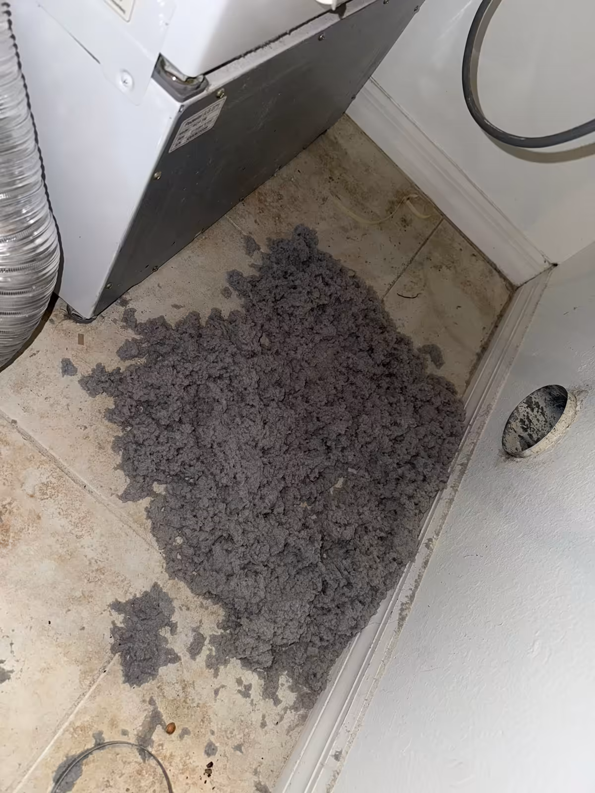 Grey lint pulled from a dryer vent, piled on the laundry room floor between the dryer and the round wall vent opening