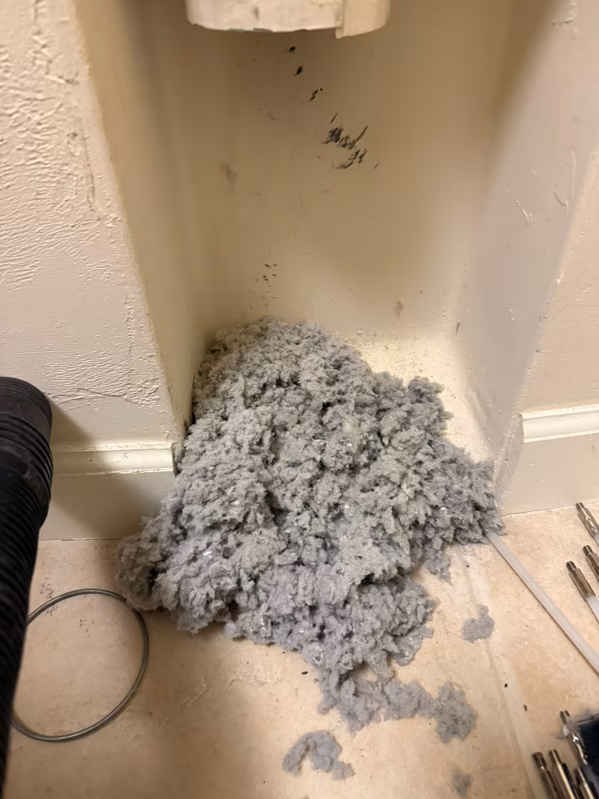 Grey lint cleared from the dryer vent line, heaped on the tile floor of a laundry alcove beside the vacuum hose and brush rods