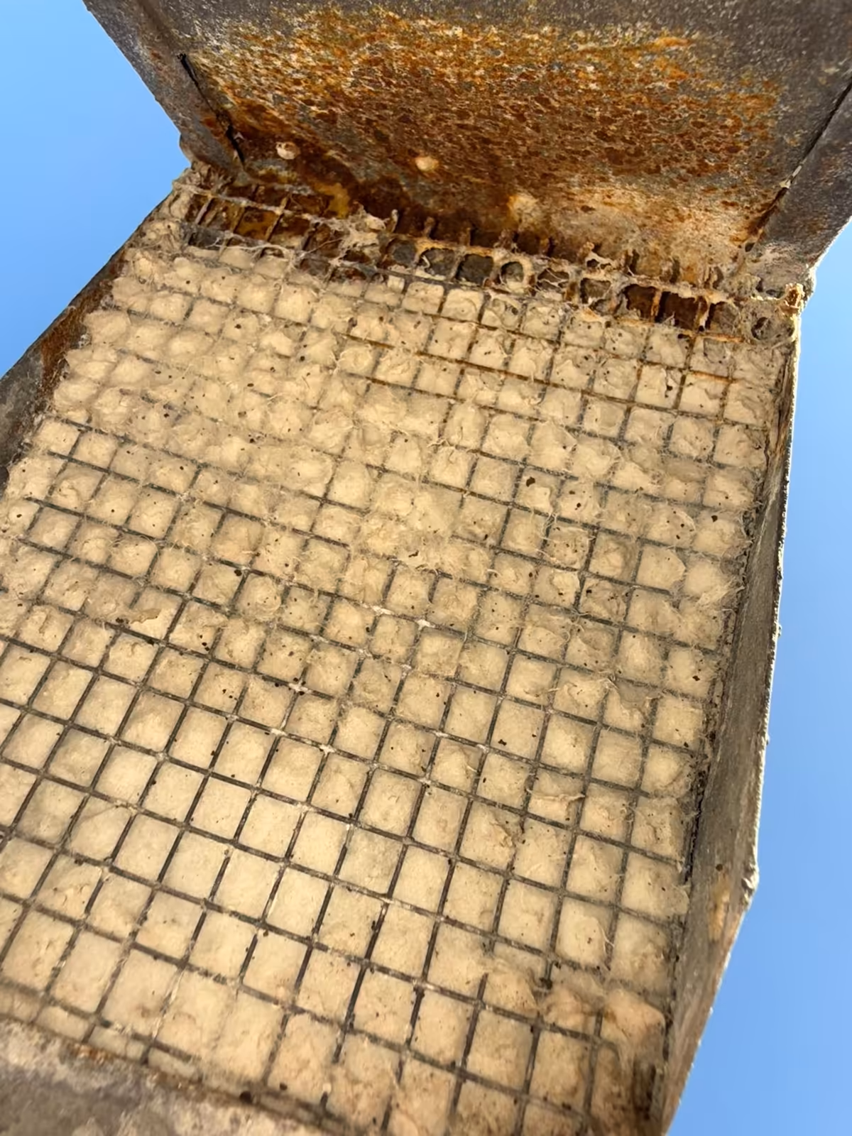 Underside of a rusted rooftop dryer vent hood, its wire screen completely blinded over with packed lint