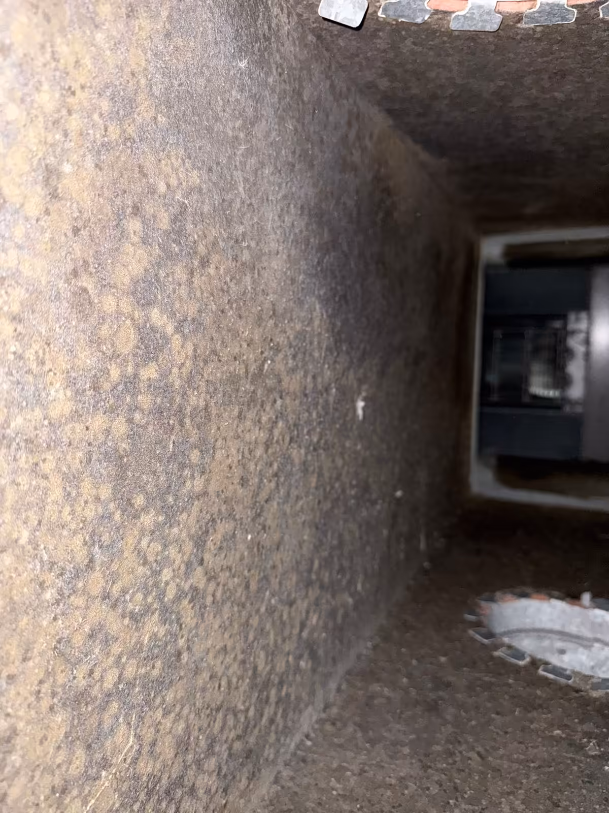 Inside a fiberglass duct board plenum before cleaning, the lining darkened brown and blotched with buildup, looking down the run toward the air handler opening