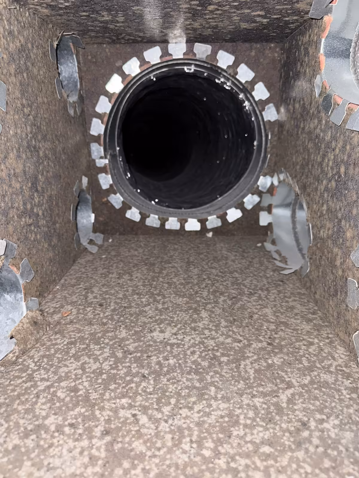 Inside the same duct board box before cleaning, dirt speckled across the walls and floor around a round flexible takeoff and four cut side collars